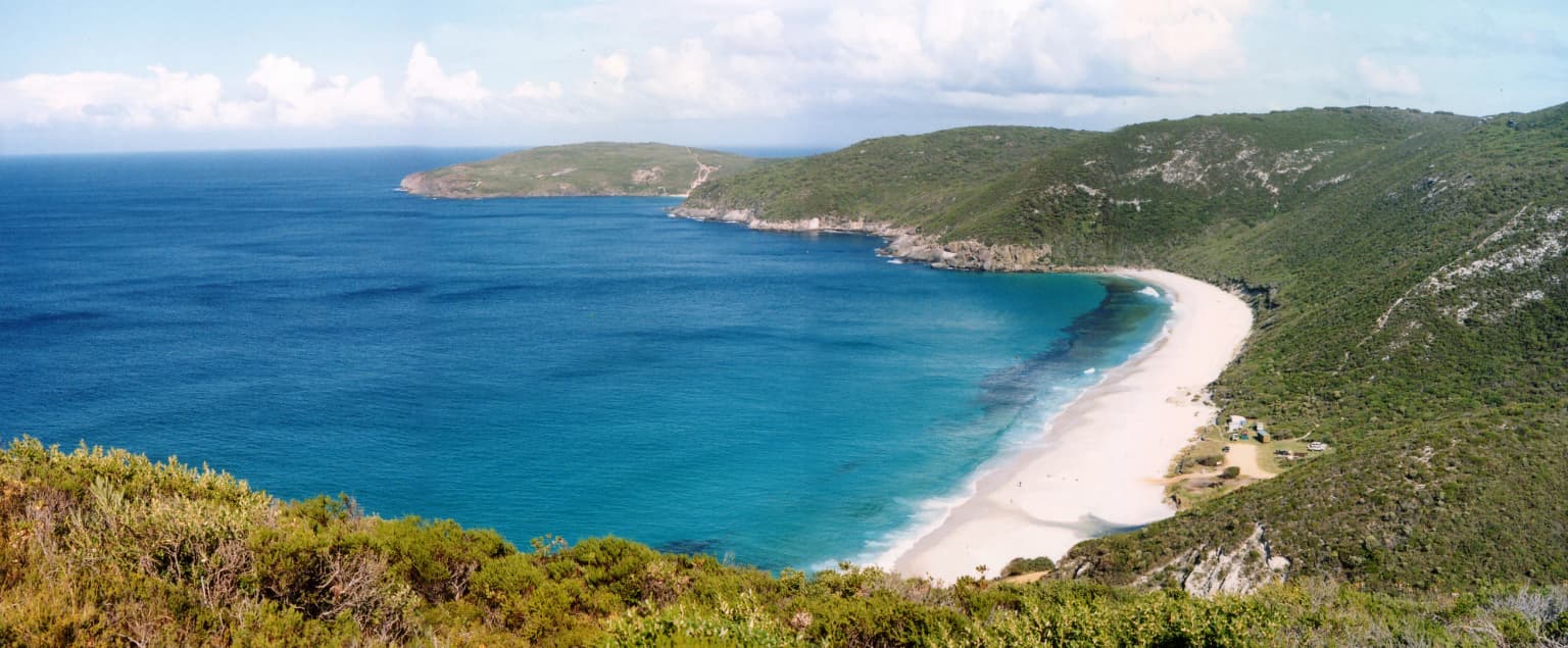 Paisaje costero con océano turquesa, playa de arena blanca, colinas verdes y promontorio rocoso