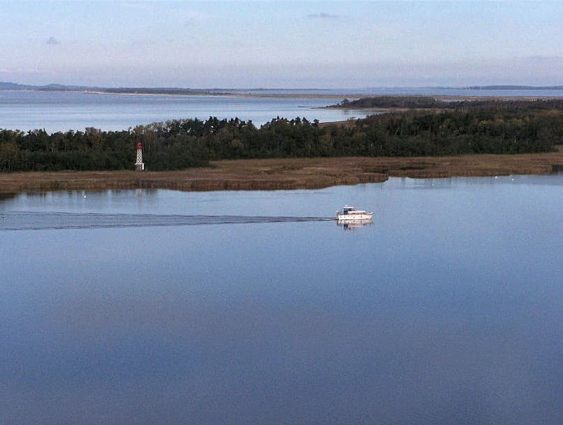 A white boat on calm blue water with a lighthouse on a distant shore and forested coastline