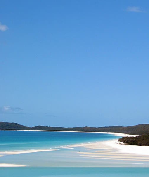 Playa de arena blanca con agua turquesa, colinas distantes bajo un cielo azul despejado