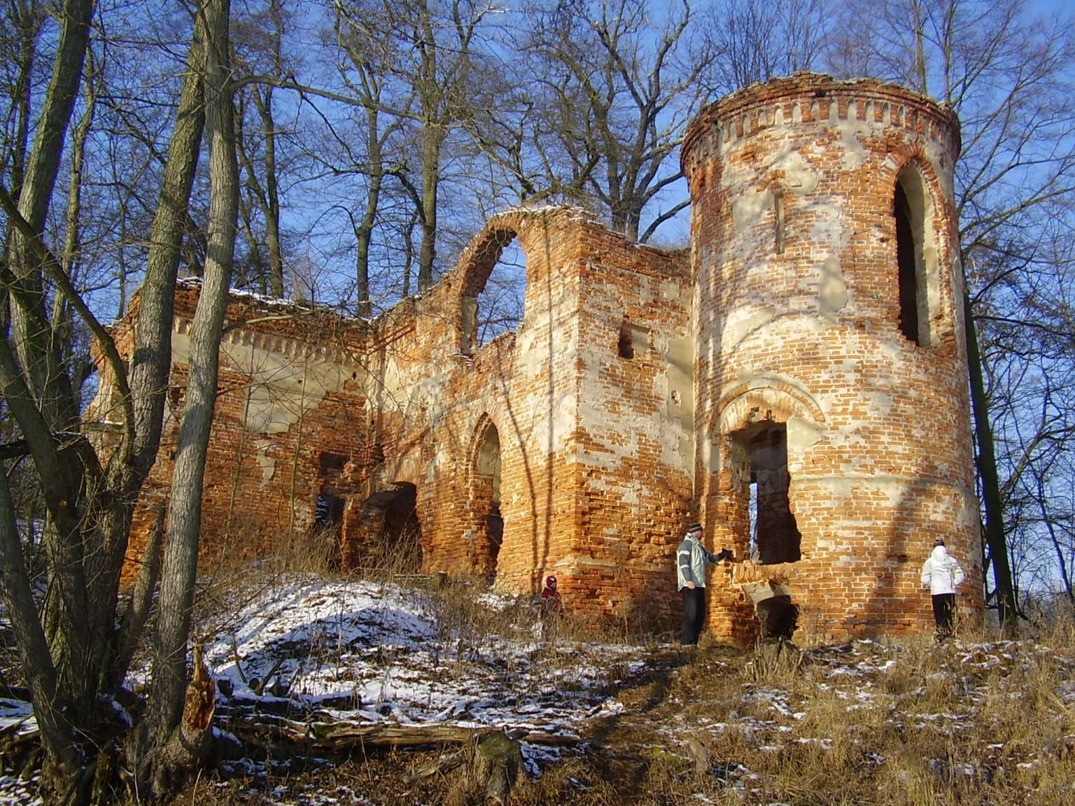 Brick castle ruins with arched openings and a cylindrical tower, surrounded by snow and trees, with two people standing nearby