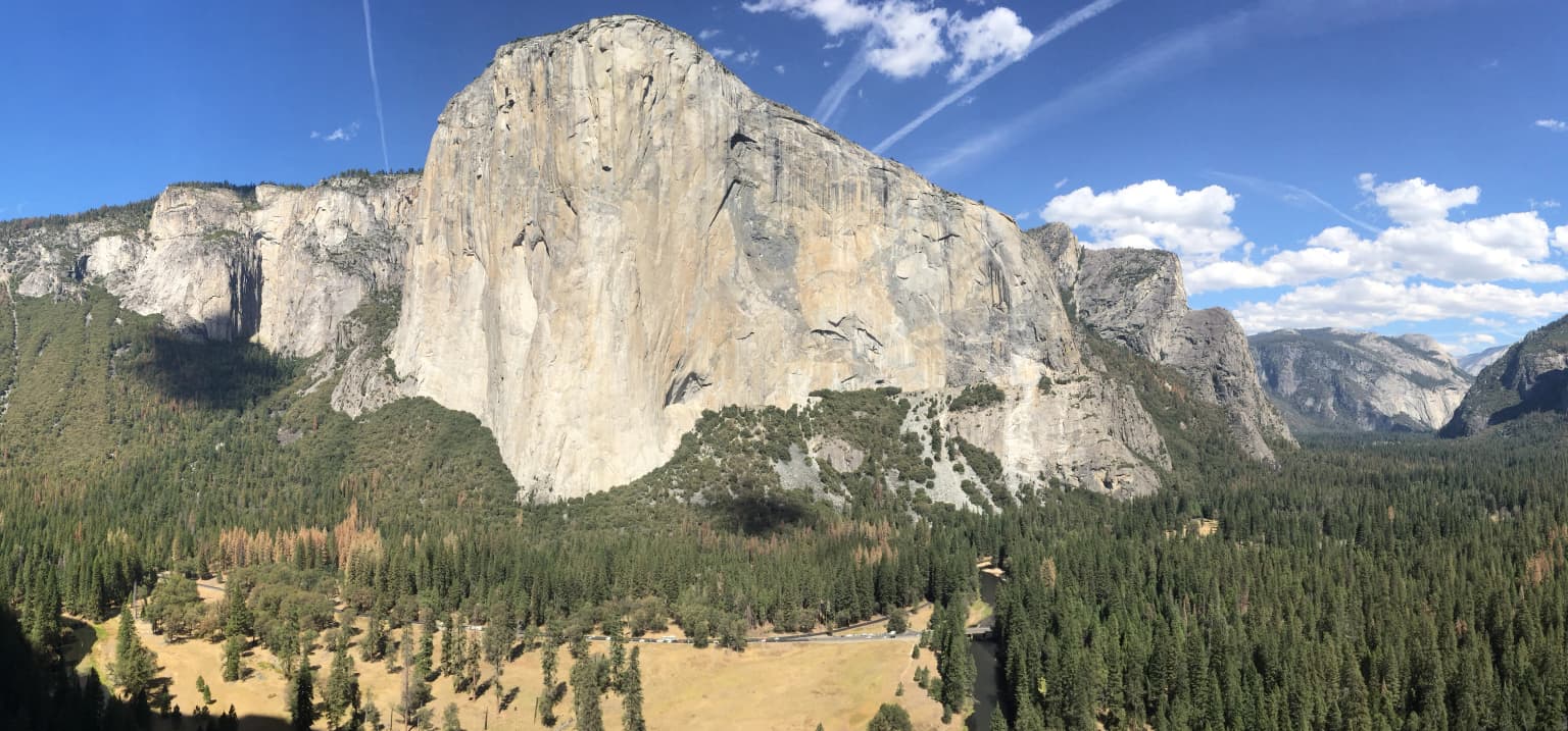 Amplia vista panorámica de la formación rocosa El Capitán en el Valle de Yosemite, con colinas boscosas y cielo azul despejado.