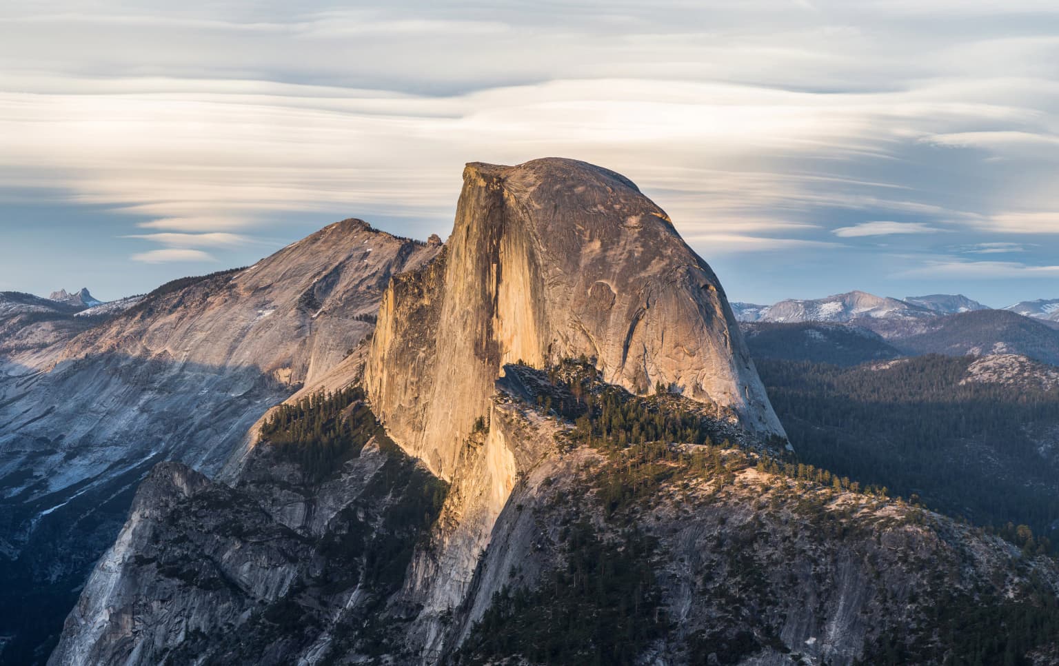 Una gran montaña con forma de domo de granito, con acantilados escarpados y una cima plana, rodeada de picos rocosos y zonas boscosas bajo un cielo parcialmente nublado