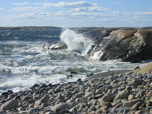 Ocean waves crashing against rocky shore with pebbly beach under partly cloudy sky