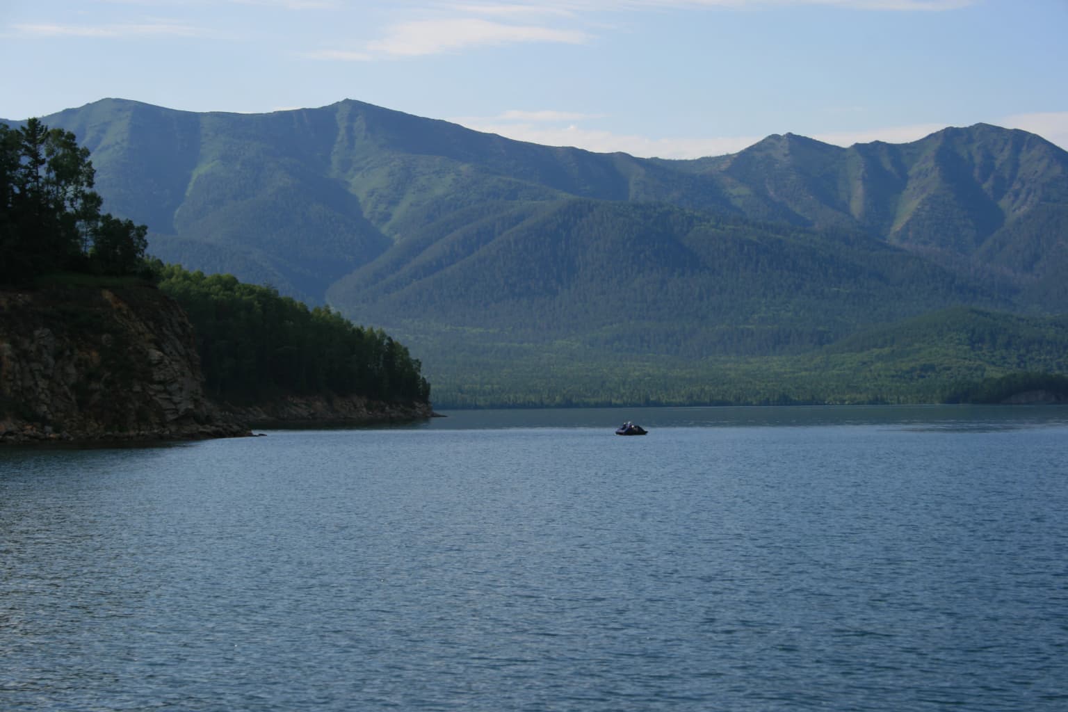 Calm blue lake water with a forested shoreline and mountain range under a clear sky