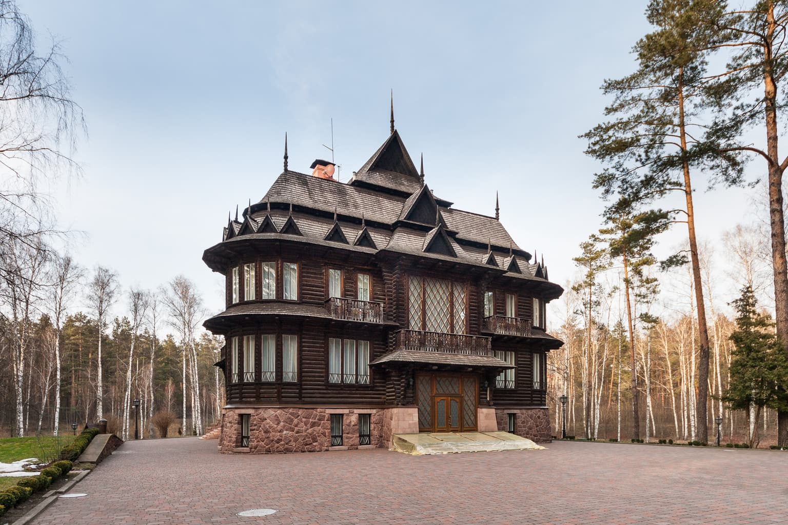 Large wooden mansion with stone foundation, steep roof, and decorative architectural details, surrounded by forest and paved driveway