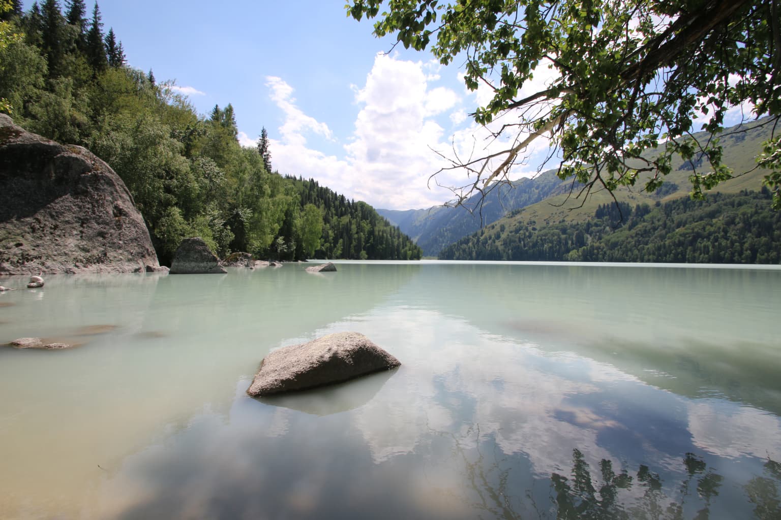 Calm lake with light blue water reflecting sky and clouds, bordered by green forested hills and rocky formations under a partly cloudy sky.