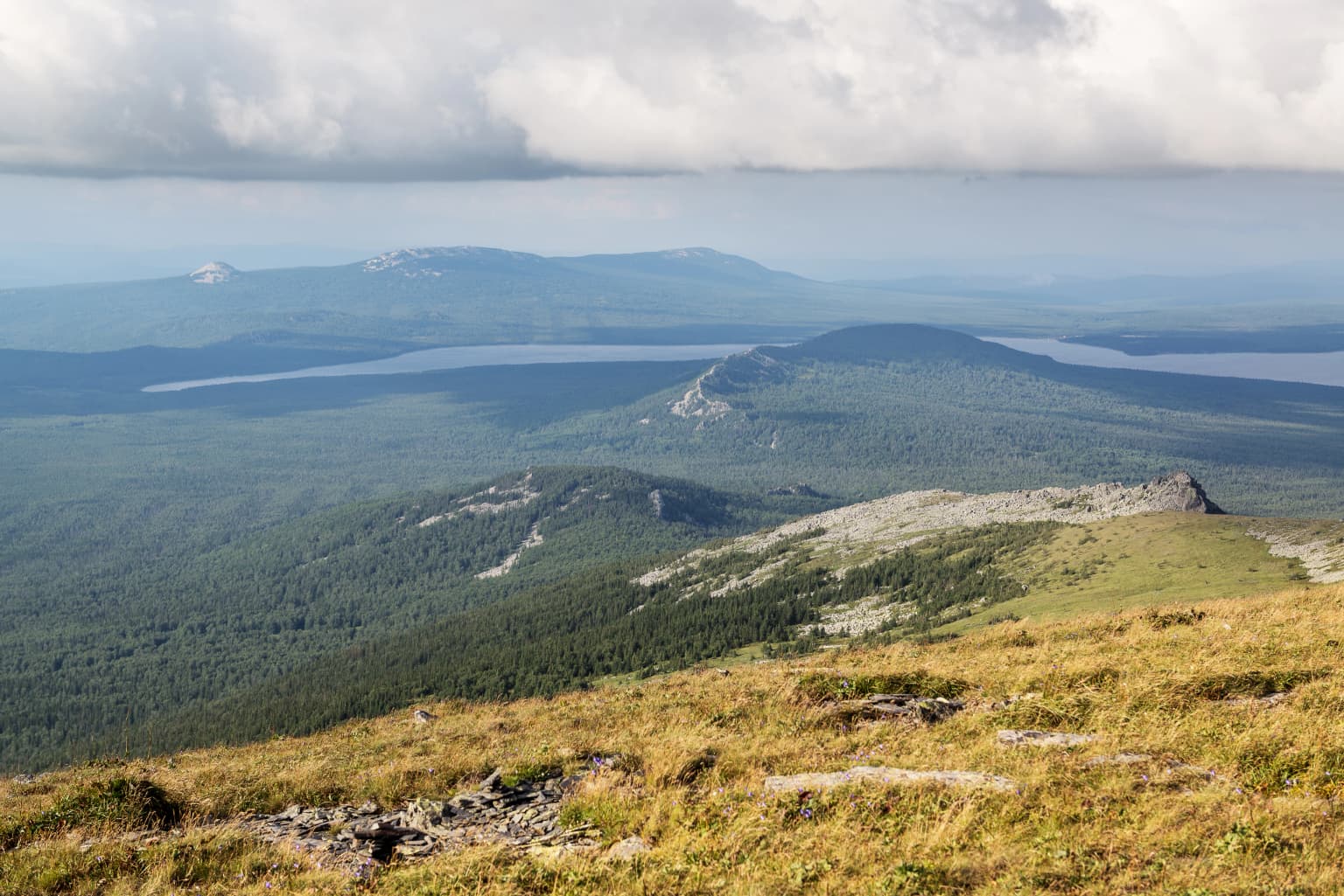 Panoramic landscape view of Zyuratkul National Park with grassy hills, forests, mountain ranges, and a lake in the distance under a cloudy sky